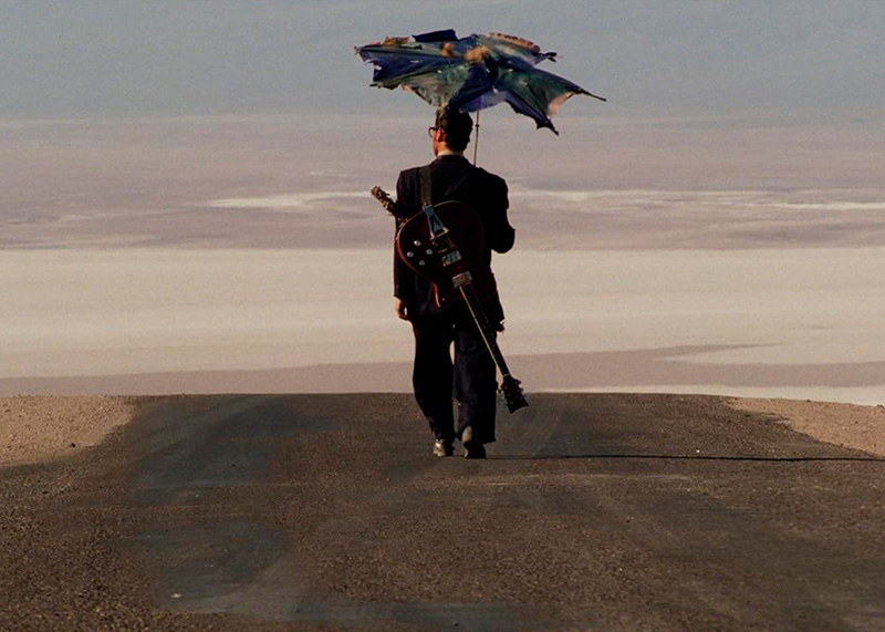 Hombre camina por una carretera solitaria sosteniendo un paraguas y cargando una guitarra y una espada.