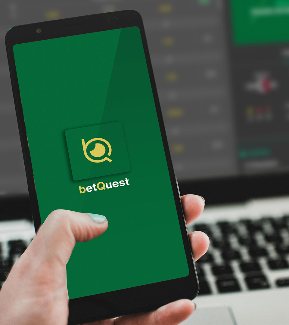 Persona con un smartphone utilizando la aplicación betquest frente a un portátil con la misma aplicación en pantalla.